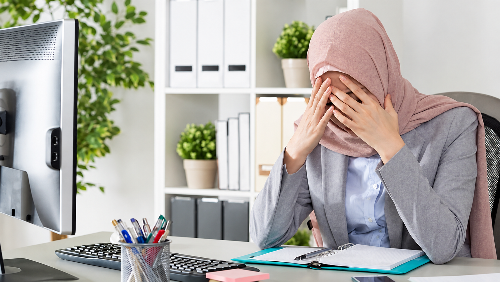 Woman wearing a dusty-rose hijab seated at a desk, covering her face with her hands