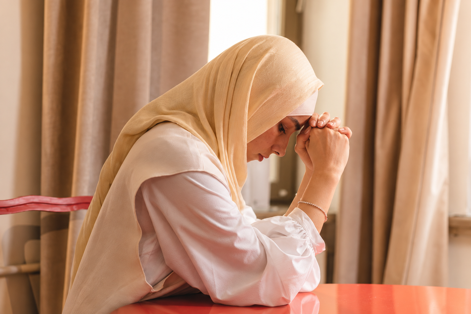 Woman in a beige hijab resting her head on her hands in a warmly lit room